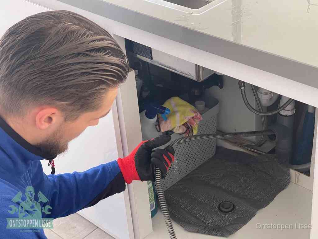 Een monteur voert een flexibele ontstoppingsveer in onder een keukenkastje om een verstopping voorbij de sifon te bereiken in Lisse