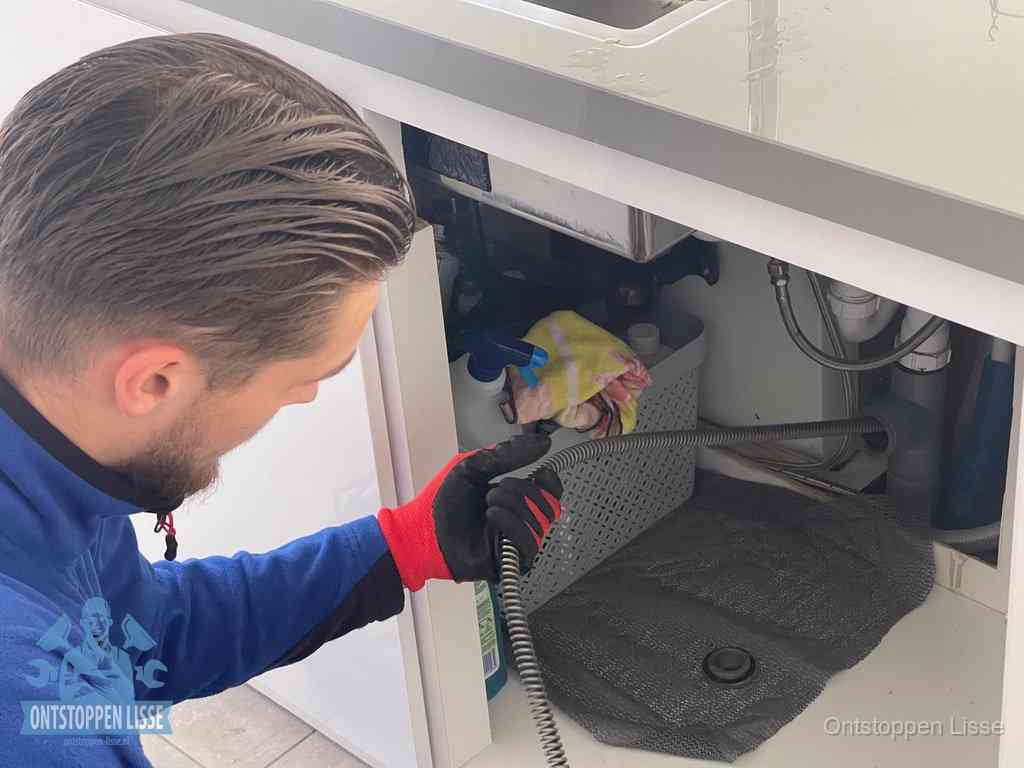Een monteur voert een flexibele ontstoppingsveer in onder het keukenkastje, langs de sifon, om de gootsteen te ontstoppen in Lisse