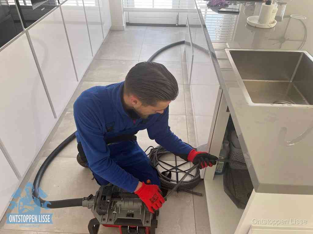 Een monteur ontstopt de keukenafvoer onder de spoelbak met een elektrische veermachine in Lisse