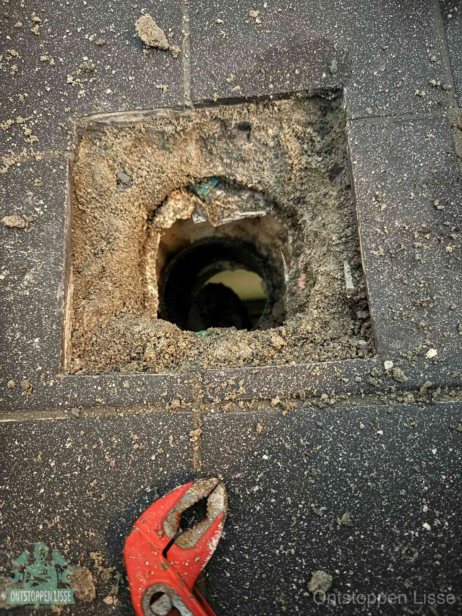 Een geopend rioolputje in een betontegelvloer met losgemaakt rooster en een zichtbare ronde afvoerbuis, het achterhalen waar de afvoerverstopping zit in Lisse