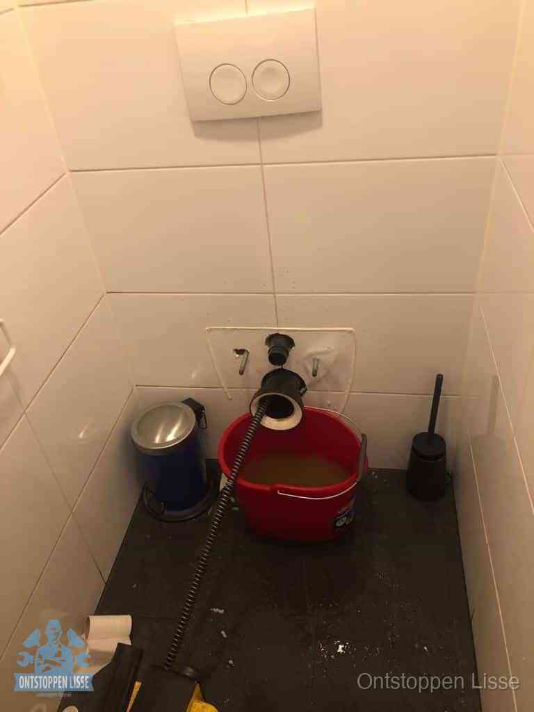 Een ontstopte wand-wc met verwijderde toiletpot waar een ontstoppingsveer in de wanduitgang verdwijnt, het achterhalen waar de verstopping zit in Lisse
