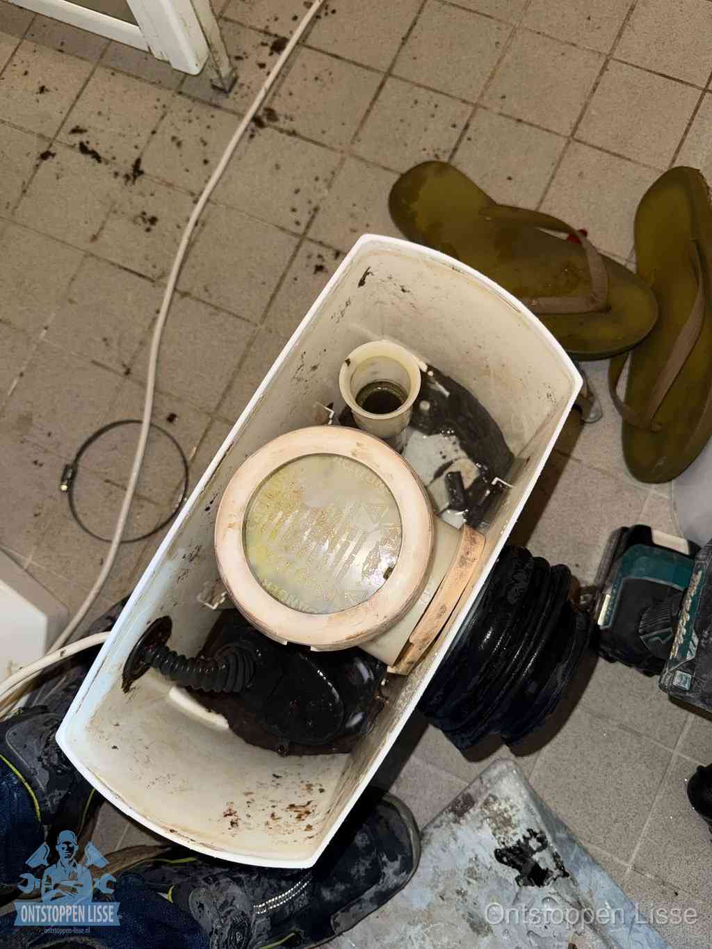 Een gedemonteerde toiletpot op de vloer met ontstoppingsveer en gereedschap ernaast, het verwijderen van een vastzittend voorwerp uit de wc in Lisse