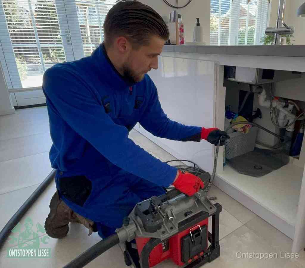 Een monteur met rode handschoenen knielt in een keuken en voert een elektrische ontstoppingsveer in tijdens het professioneel afvoer ontstoppen in Lisse